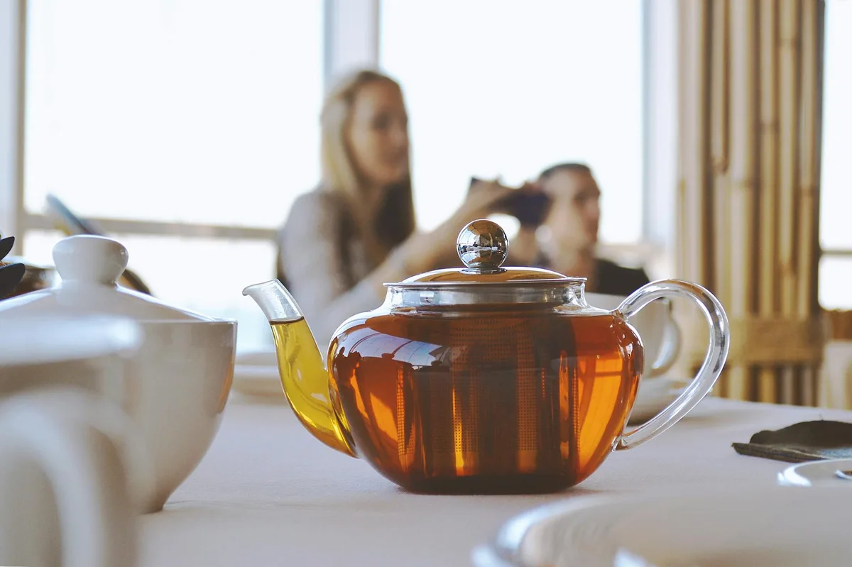 A warm close-up of a glass teapot with hot tea on a restaurant table during the day.